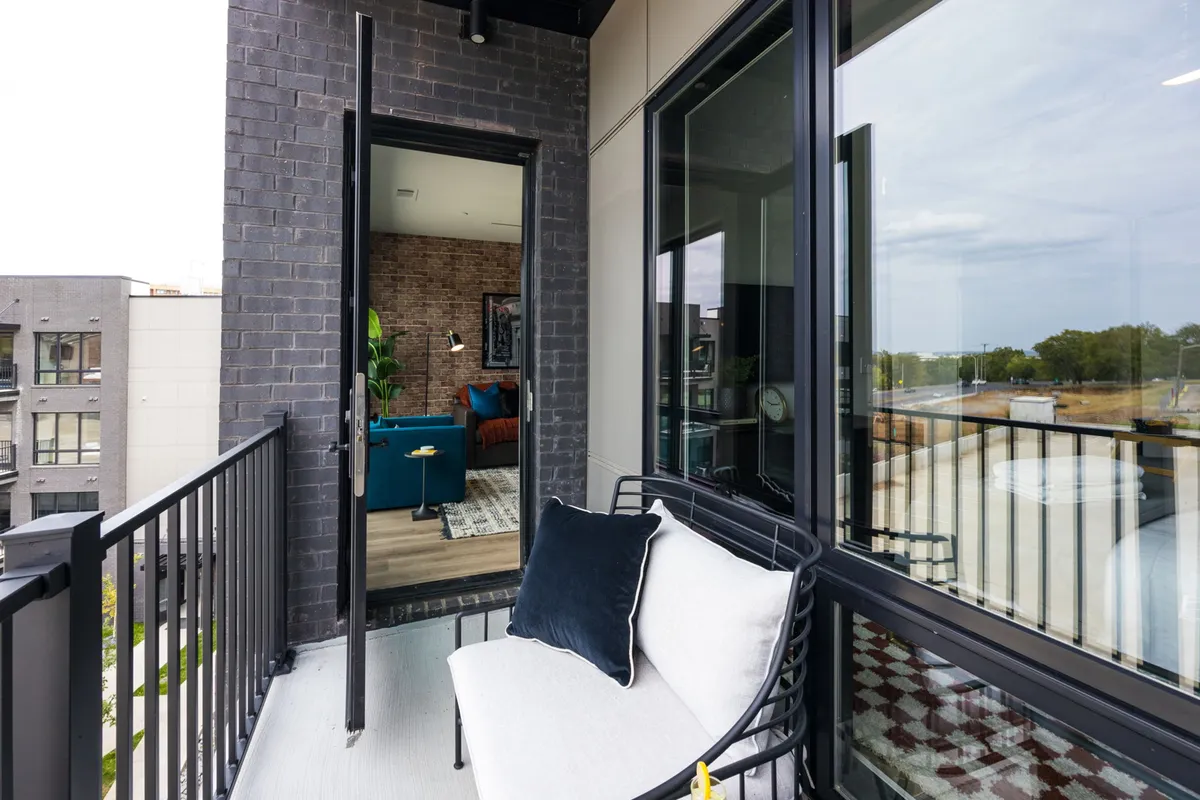 This exterior shot shows a stylish balcony area with seating, a green plant, and view into the condo's interior through glass doors. The checkered floor and decorative lamp add to the inviting, urban vibe.