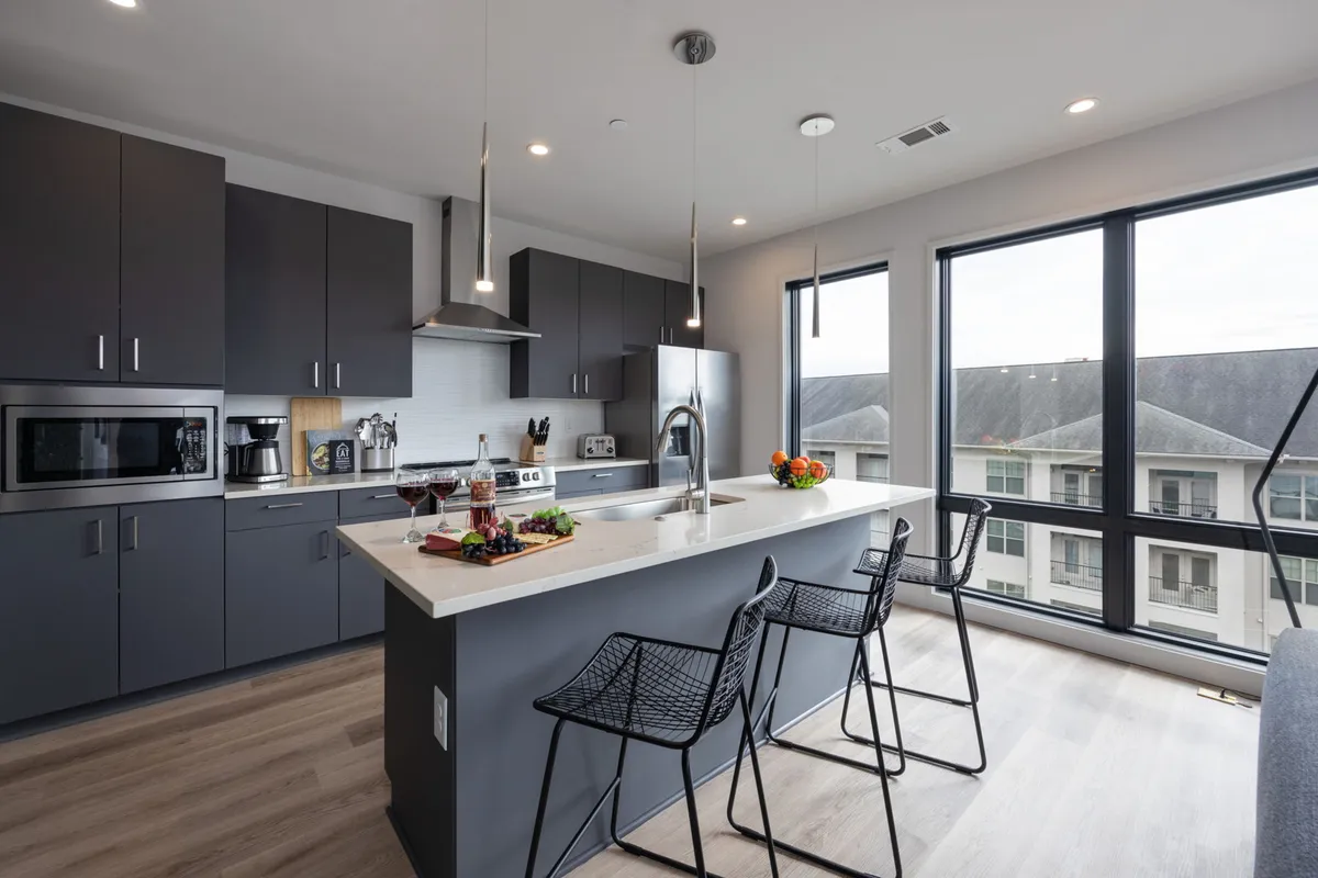 A contemporary kitchen with a large island, bar stools, fresh fruit arrangement, wine glasses, and expansive windows letting in natural light. The scene is set for socializing, cooking, and shared meals, reinforcing a sense of community.