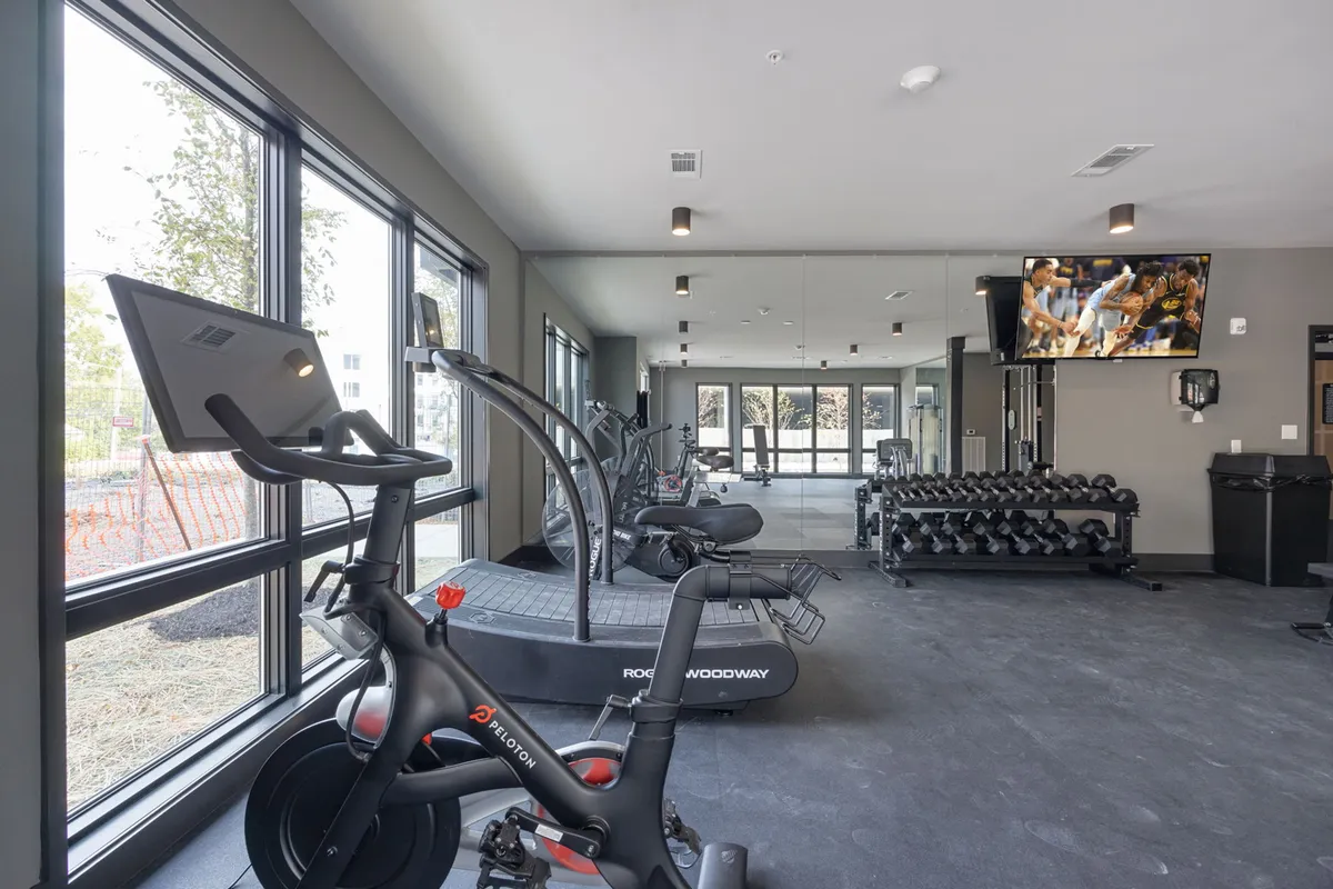 Inside the building's gym, a Peloton bike, treadmill, dumbbell rack, and television screen are showcased. Large windows let in ample natural light, creating a bright, motivational space for workouts and personal fitness routines.