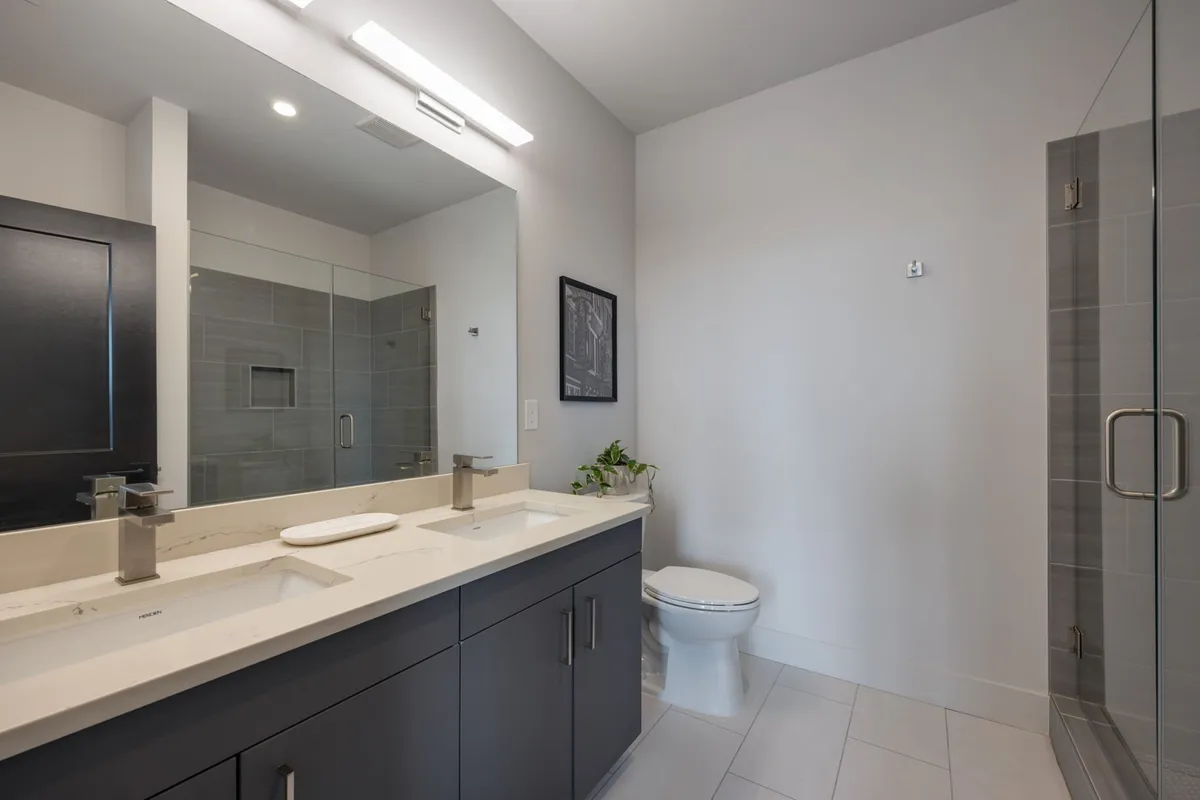 A serene and spacious en-suite bathroom featuring dual sinks, a large mirror, a glass shower, and a calm, modern design. The bathroom is filled with soft daylight, creating a tranquil space for personal routines and relaxation.