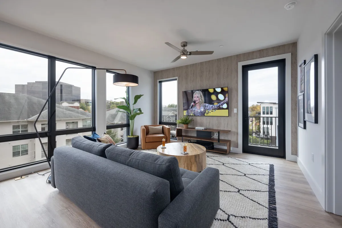 A contemporary living room featuring a plush sofa, coffee table, television, potted plant, and large windows that offer natural light and glimpses of the city outside. The setup is ideal for watching TV, entertaining, or casual conversations.