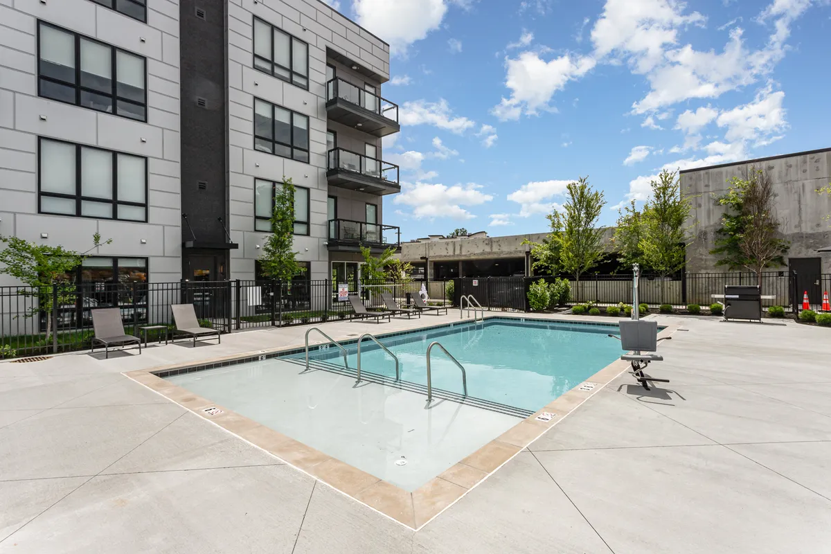 This image features a sparkling swimming pool with an adjacent balcony area surrounded by lush greenery. The scene captures a relaxed atmosphere with inviting poolside seating and views of the building's exterior amenities.