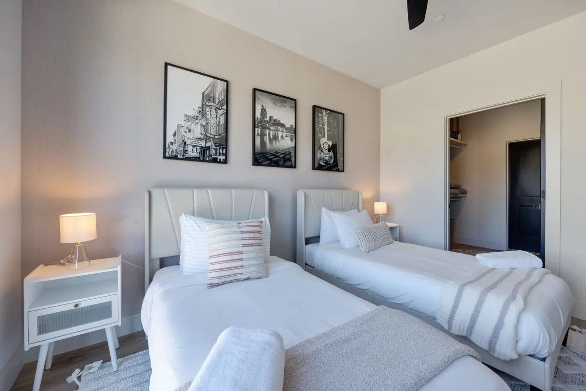 A tranquil second bedroom setup with two twin beds, soft bedding, framed black and white photographs above the beds, and nightstands with lamps. The room is filled with peaceful, natural light, offering flexibility for families, friends, or colleagues sharing the space.